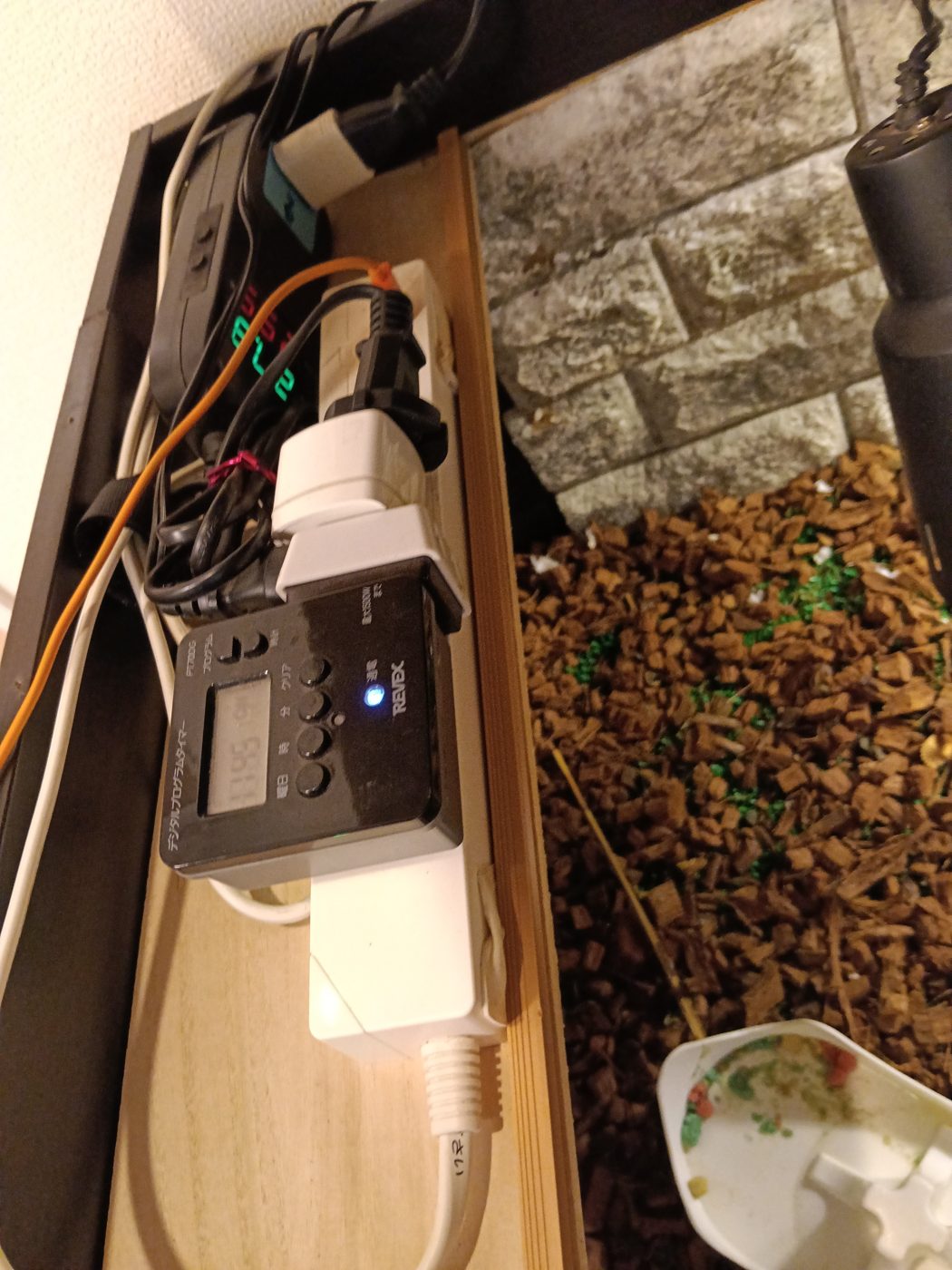 リクガメケージの側板に固定した電源タップとREVEXのデジタルプログラムタイマー、奥にサーモスタットの表示 - UVBライトのタイマー管理